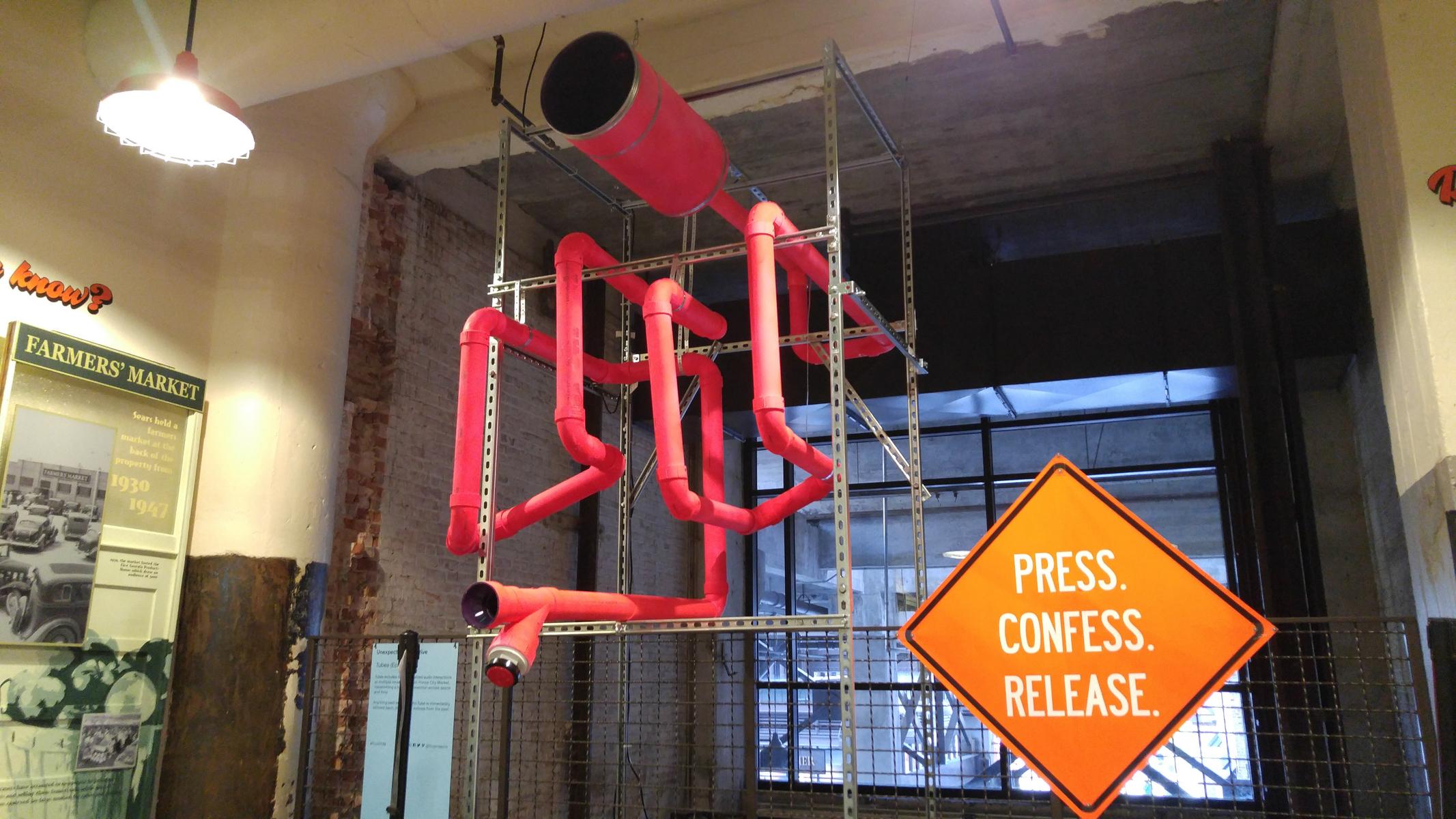 A photograph of the Echo Tube interactive sound installation: a series of fluorescent pink tubes with a red button attached. A construction-style sign instructs visitors to 'Press. Confess. Release.'