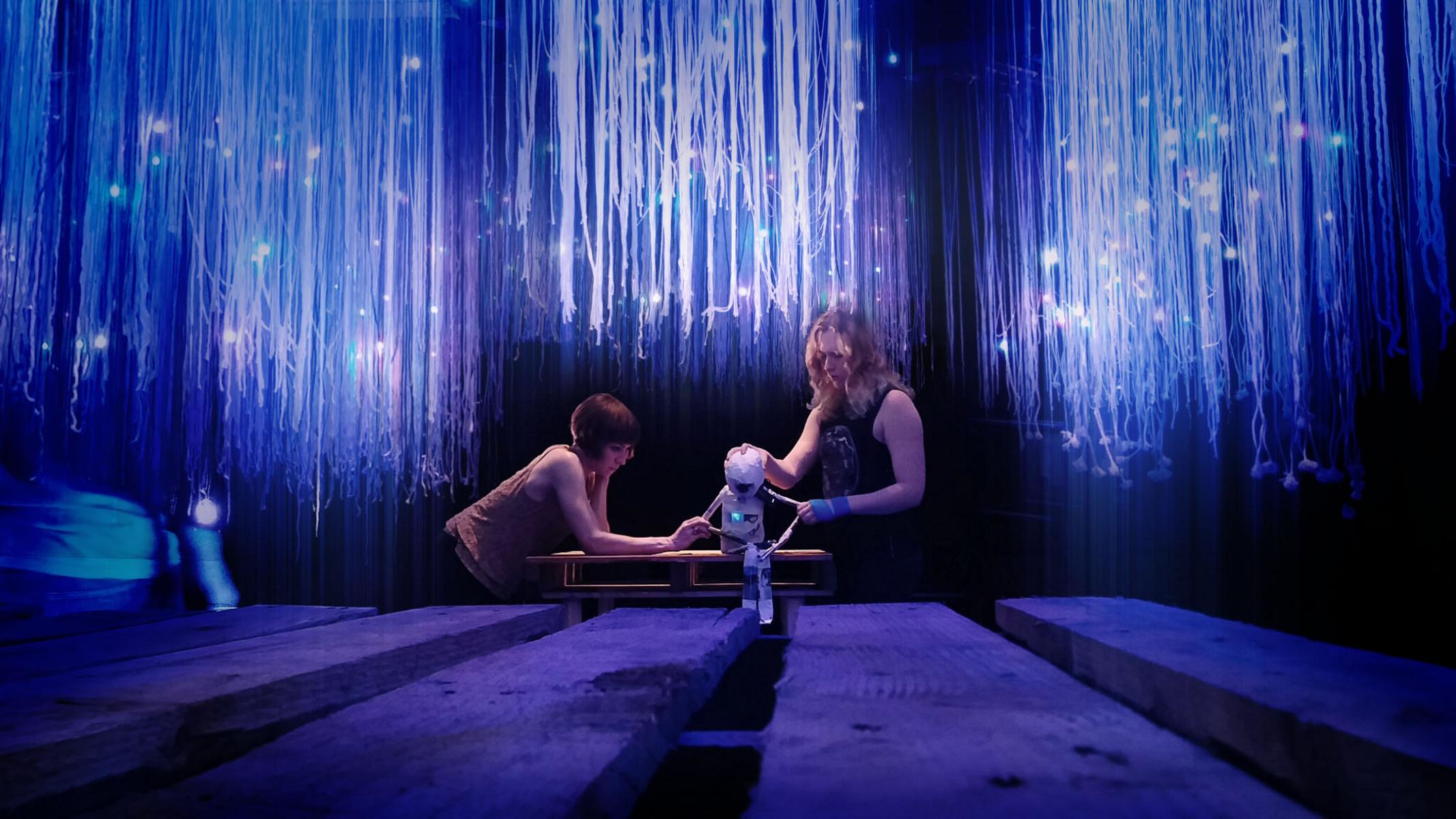 Two puppeteers rehearse with a white puppet seated on a shipping pallet table. Suspended above are shipping pallets from which string and points of light hang.